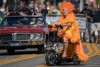 A drag queen dressed in orange rides a scooter down the parade route