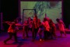 A group of zombie actors reaches toward the audience under red and green lighting a projection of city streets glowing behind them